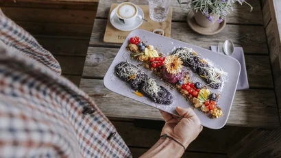 Person hält Teller mit kreativ dekoriertem Dessert und Cappuccino im Hintergrund