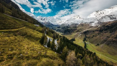 Gebirgsstraße in grüner Alpenlandschaft mit schneebedeckten Gipfeln und blauem Himmel
