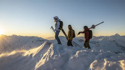 Drei Skifahrer auf schneebedecktem Berggipfel bei Sonnenuntergang