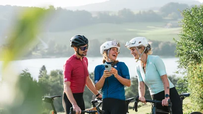 Drei Radfahrer in Sportkleidung lachen und schauen auf ein Handy am See