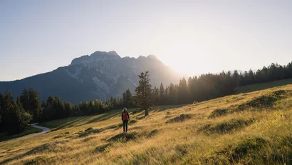 Wanderer auf sonniger Wiese vor Bergkulisse bei Sonnenuntergang