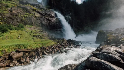 Mächtiger Wasserfall umgeben von Felsen und Wald mit zwei Wanderern auf der Wiese