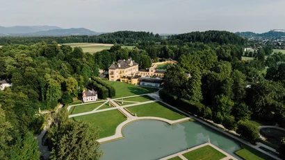 Luftaufnahme eines historischen Schlosses mit Gärten und Teich umgeben von Wäldern