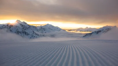 Frisch präparierte Skipiste vor verschneiten Bergen bei Sonnenaufgang