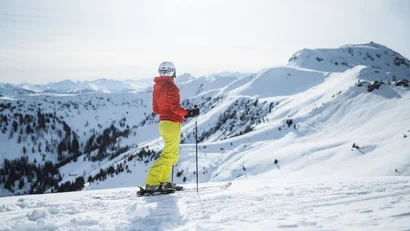 Skifahrer in roter Jacke und gelber Hose blickt auf verschneite Berge